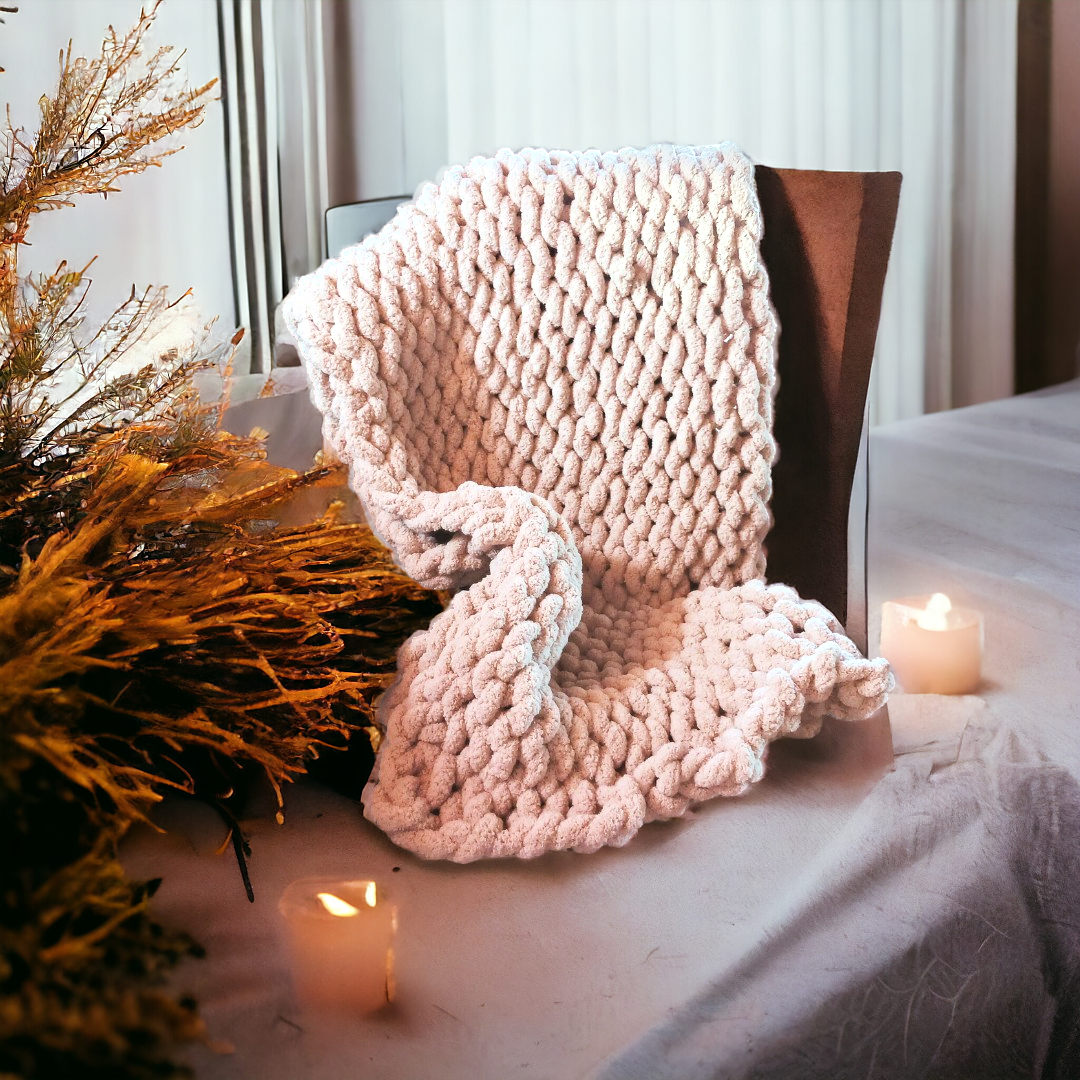 A beige chunky knit blanket draped over a chair with candles and decorative foliage in the background, depicting a cozy setting.