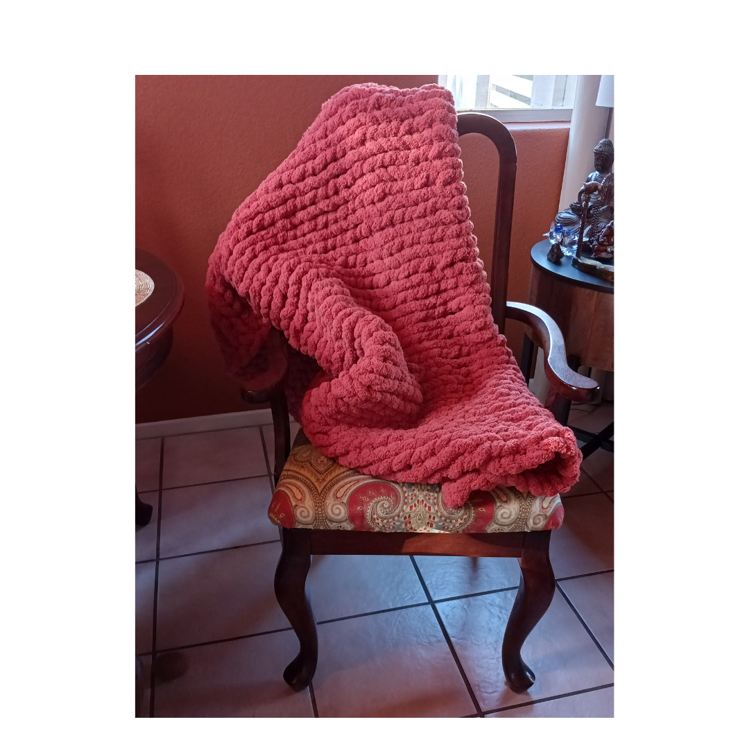 A thick, reddish-pink chunky knit blanket draped over a vintage wooden chair with an ornate, patterned cushion.