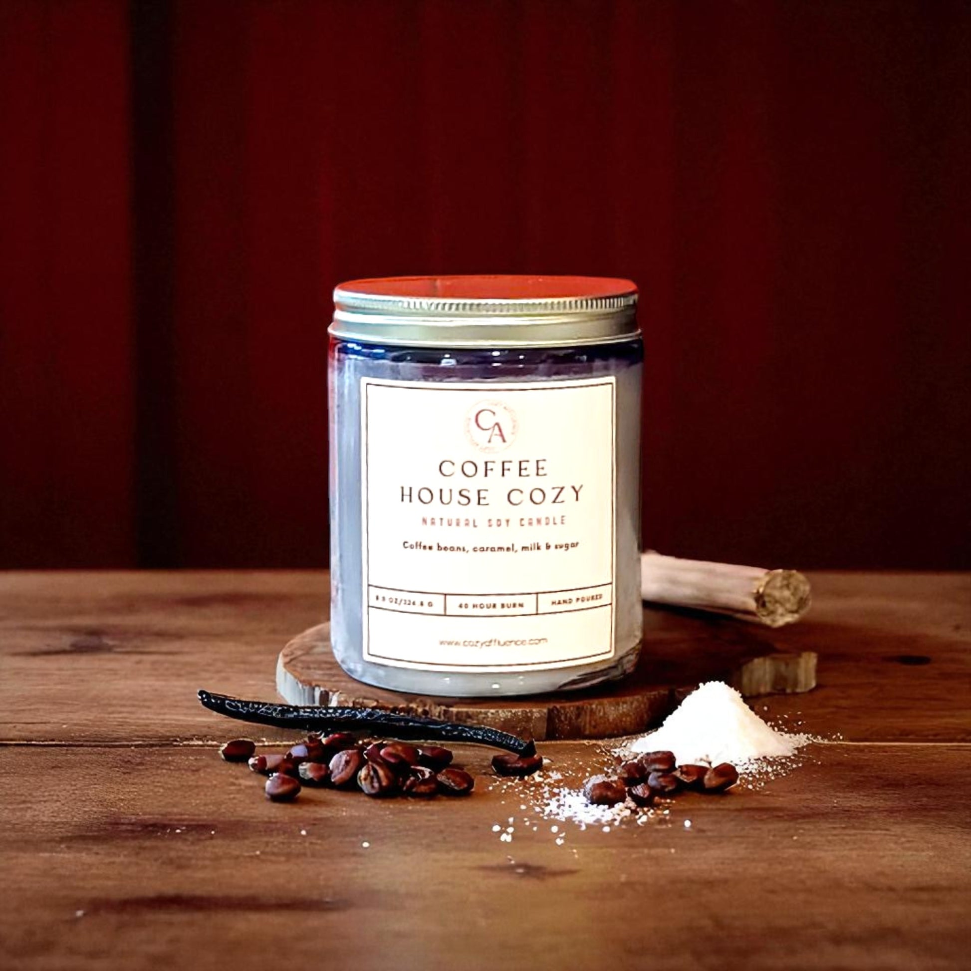 A Coffee House Cozy soy candle in a clear jar with a red lid sits on a wooden slice, surrounded by coffee beans, a vanilla bean, and a pile of white sugar.