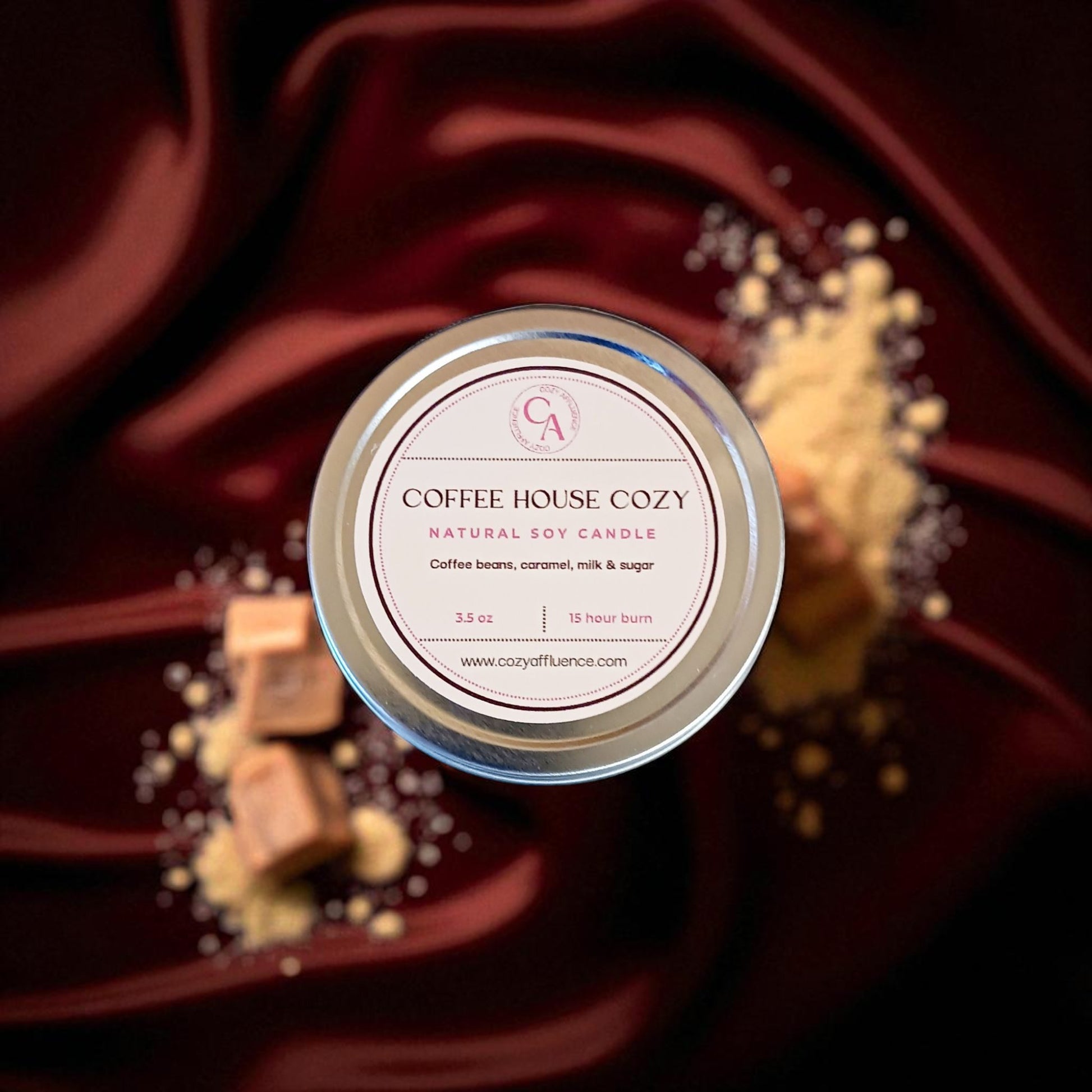 An overhead shot shows a round tin container for a Coffee House Cozy soy candle, surrounded by scattered coffee beans, chunks of caramel, and a pile of granulated sugar.