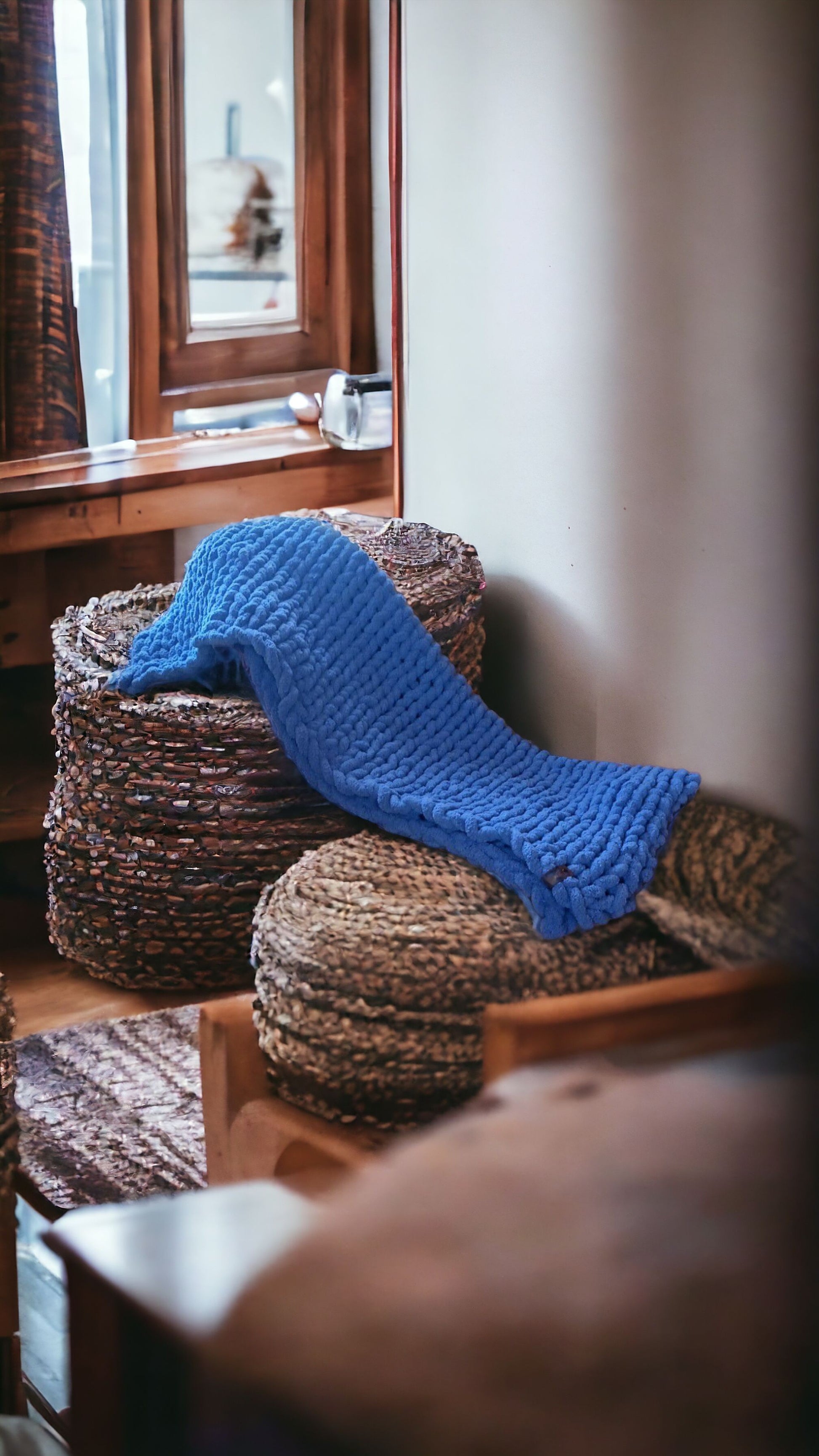 A blue chunky knit blanket draped over a woven basket on a wooden floor, with a partially visible mirror and curtain in the background.