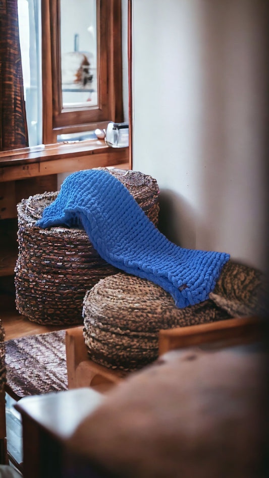 A blue chunky knit blanket draped over a woven basket on a wooden floor, with a partially visible mirror and curtain in the background.