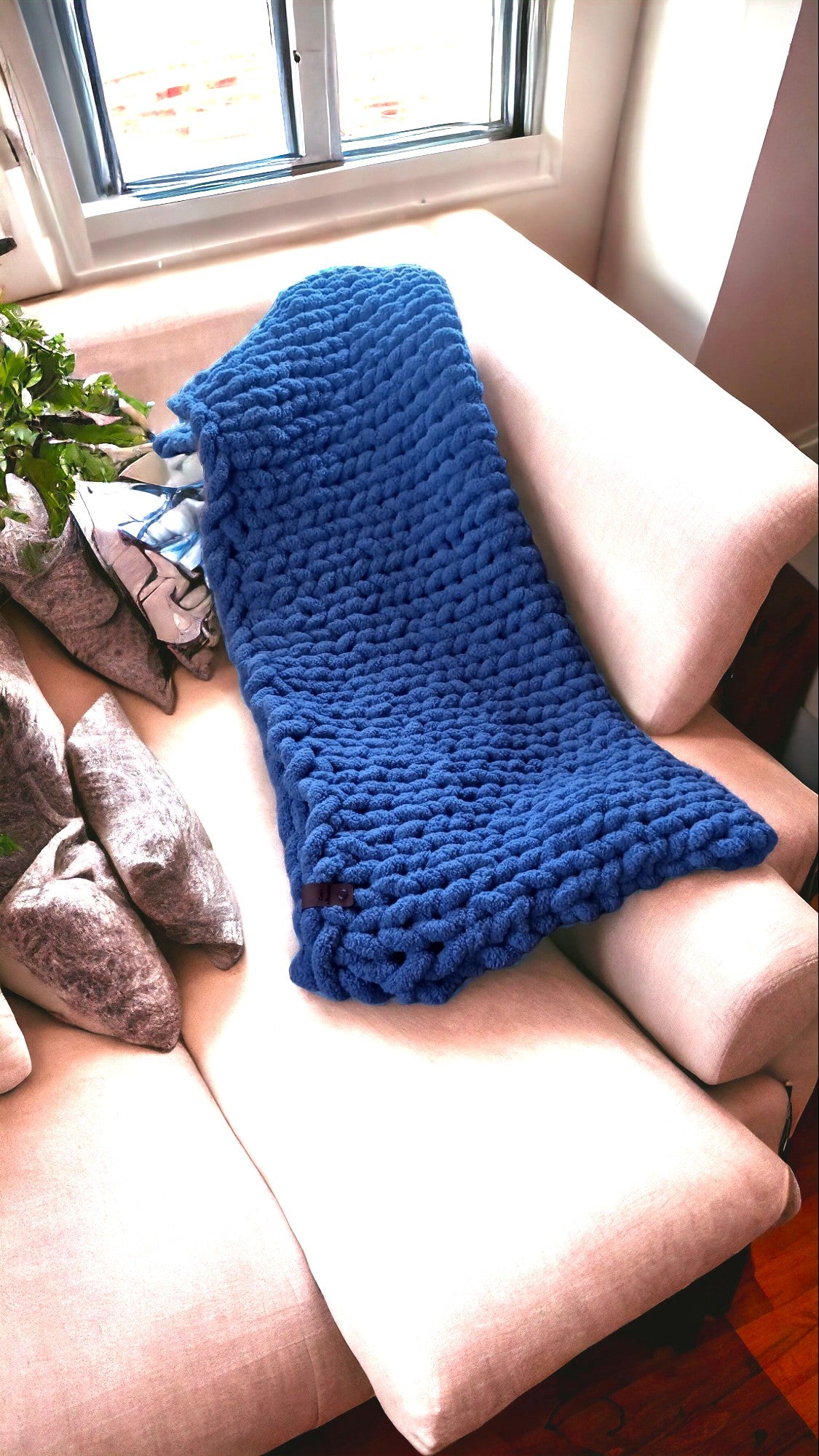 A thick, royal blue knitted blanket is folded over the back of a light-colored sofa near a window.