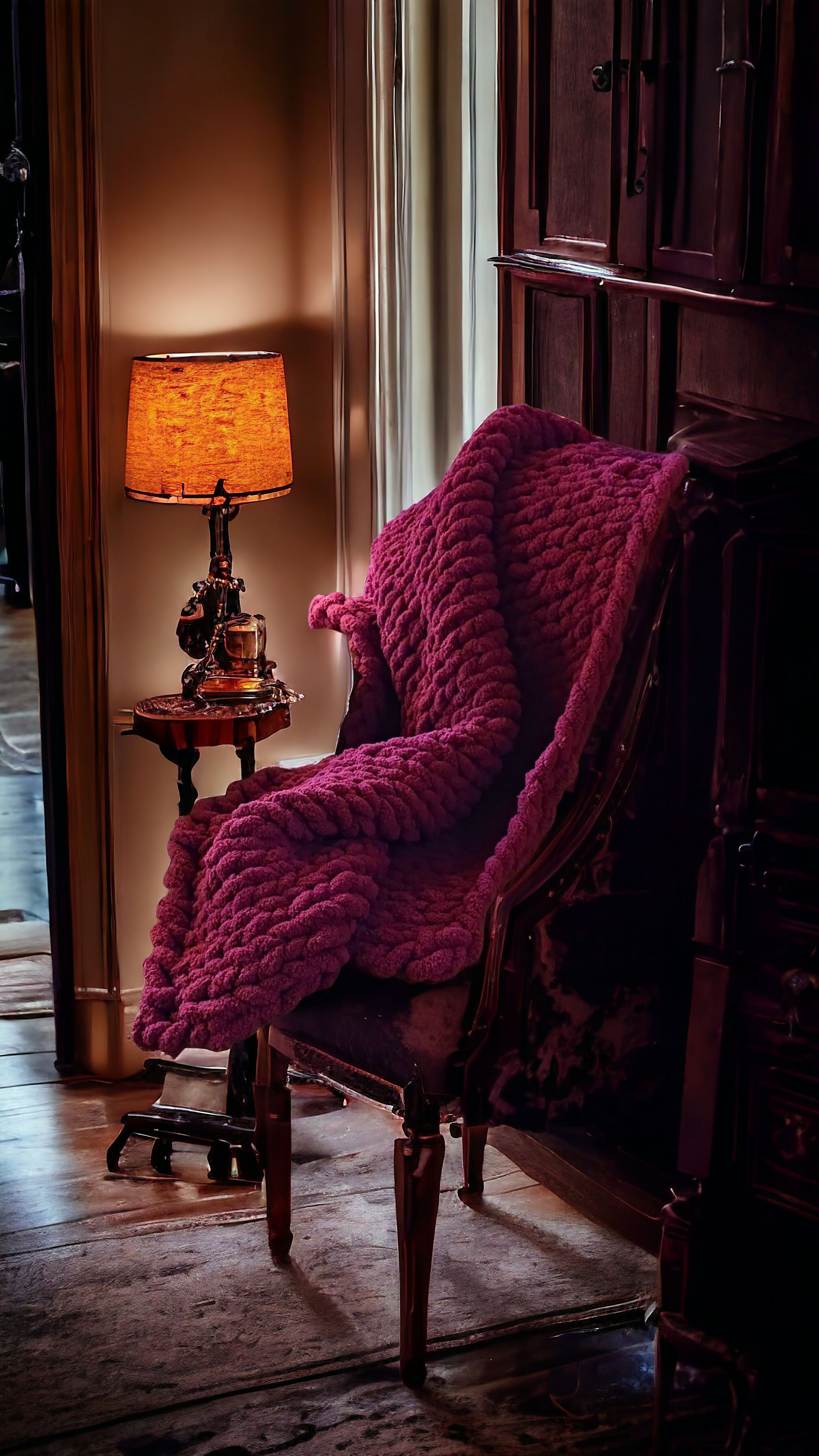 A cranberry chunky knit blanket in a deep red or cranberry color, draped over a chair next to a lamp with a warm light.