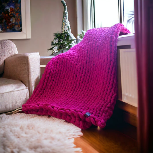 A chunky knit blanket in a bright magenta color is draped over the back of a chair, with a cozy living room setting in the background.