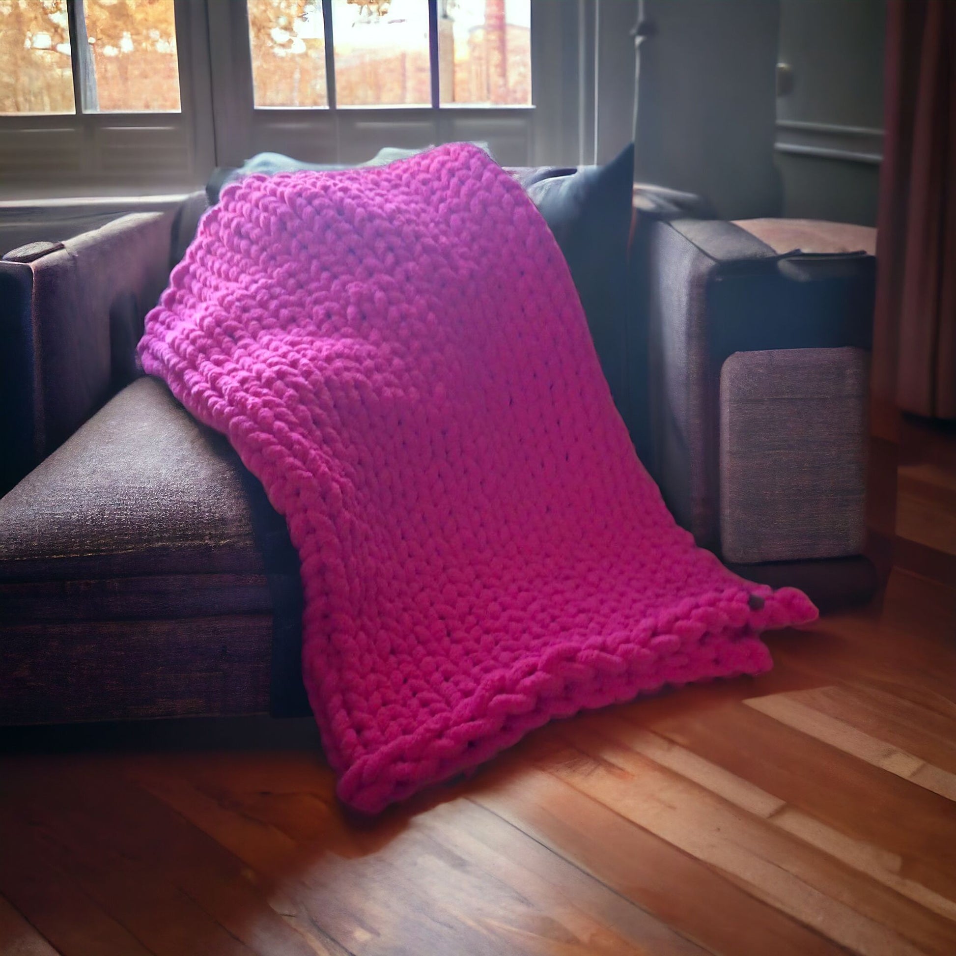 A large, vibrant magenta chunky-knit blanket is draped over a dark gray sectional sofa near a large window.
