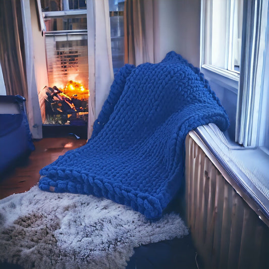 A chunky knit blanket in midnight blue draped over a chair with a cozy room and fireplace in the background.