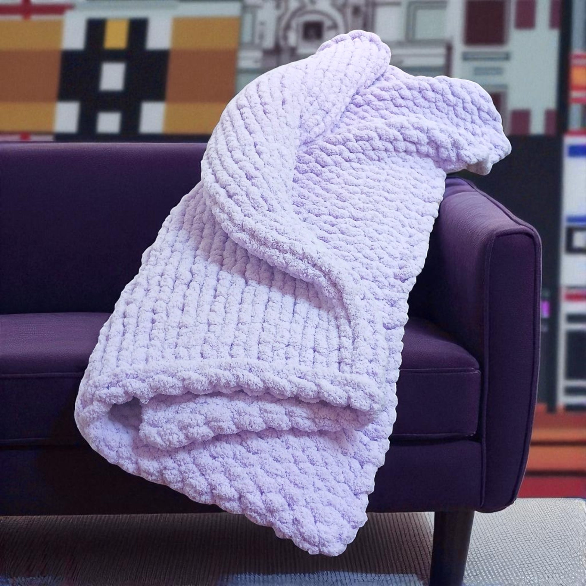 Light purple textured chunky knit blanket draped over a purple couch with a colorful abstract painting in the background.