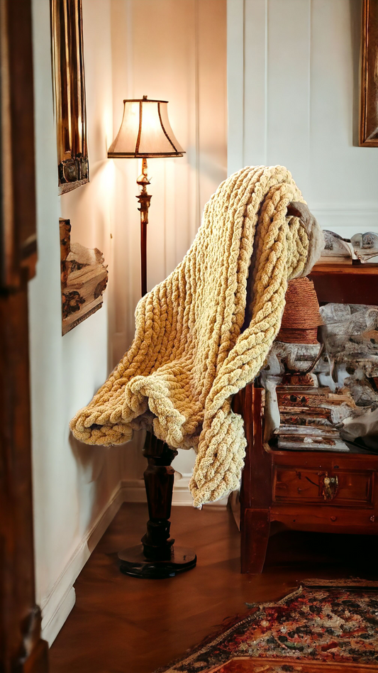 A mustard chunky knit blanket draped over the back of a chair in a room with a lamp and furniture in the background.