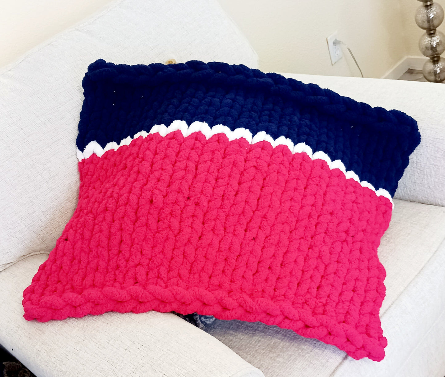 A close-up of a handmade, chunky knit blanket folded and placed on a light-colored couch, featuring a pattern of vibrant pink, a thin white stripe, and a dark blue section.