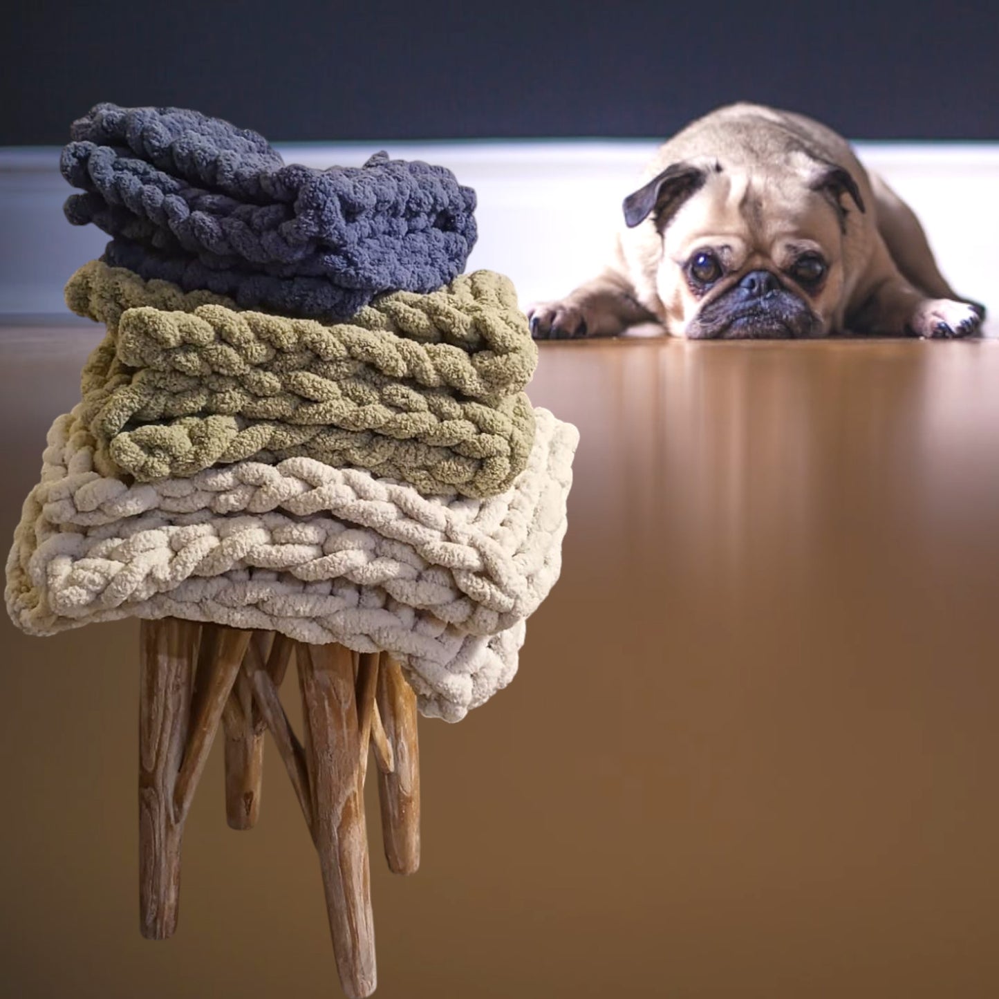 A stack of chunky knit pet blankets in beige and green colors, with a pug dog looking at them in the background.