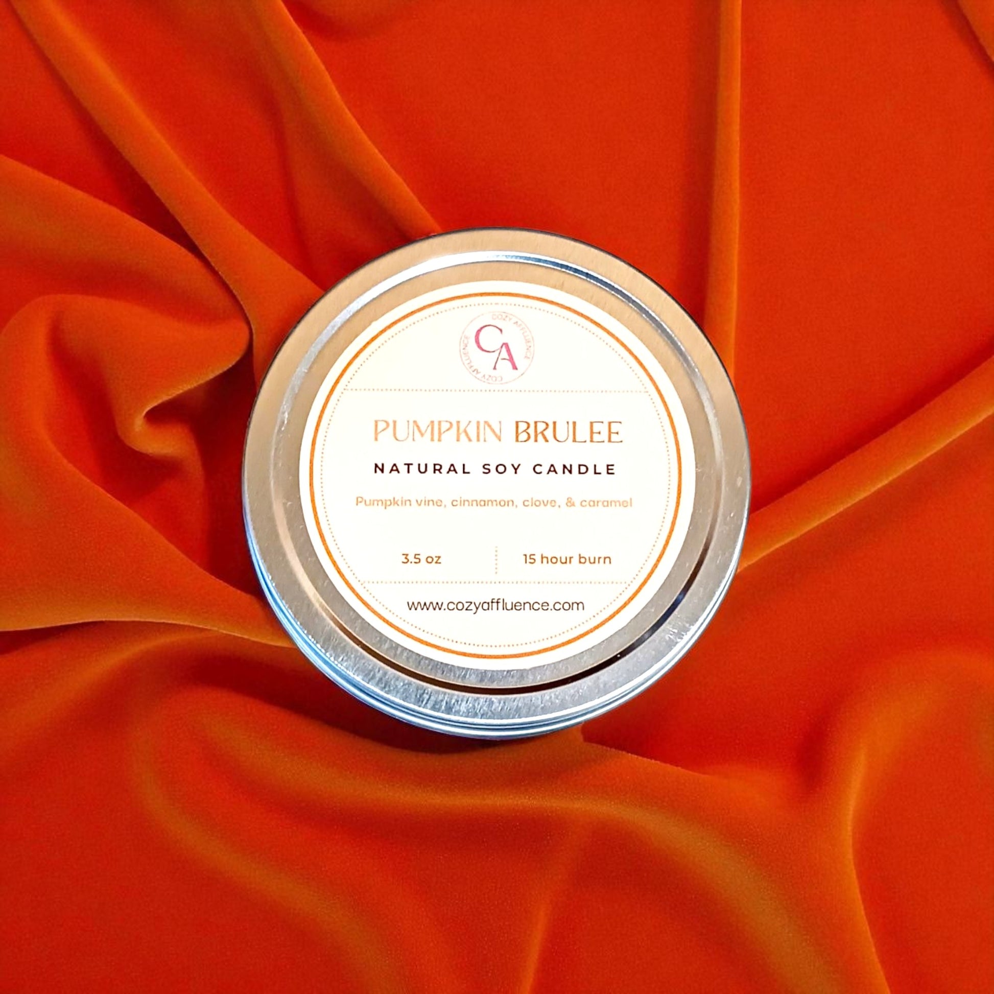 Pumpkin Brulee candle in a tin with a decorative label on an orange fabric background.