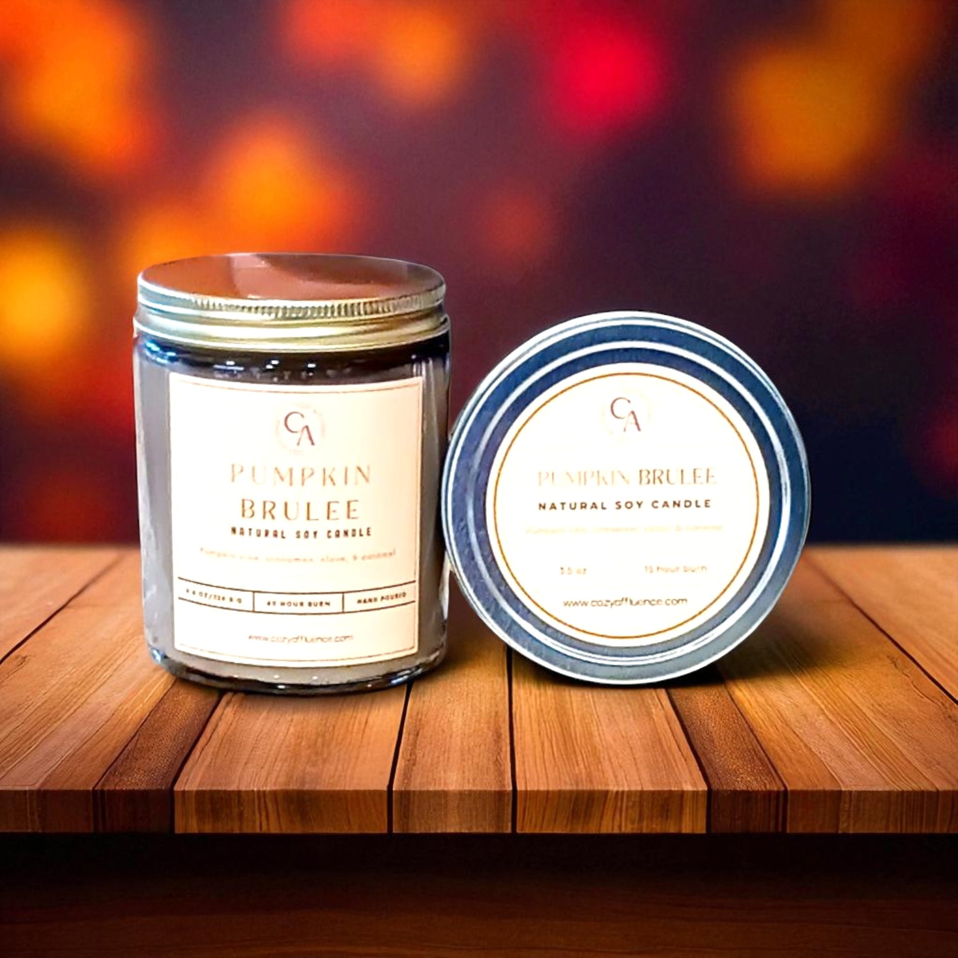 A Pumpkin Brulee candle in a glass jar with a metal lid and a matching tin container sit on a wooden surface against a blurred, warm-toned background.