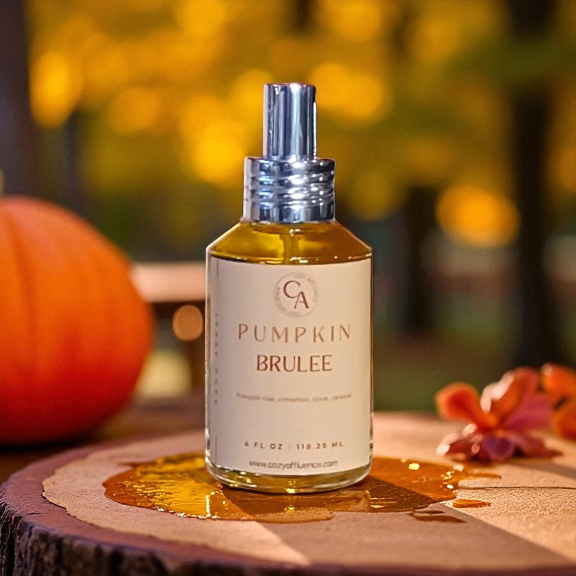 A bottle of Pumpkin Brulee room spray is shown on a wooden surface, with a pumpkin and autumnal foliage in the background.