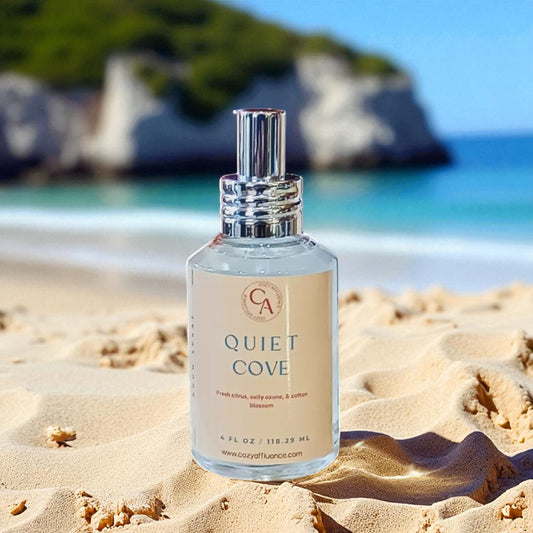 Bottle of 'Quiet Cove' room spray on a sandy beach with ocean and rocks in the background