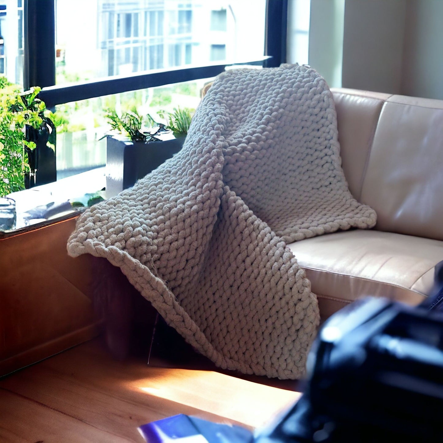 A thick, off-white chunky-knit blanket is casually draped over a cream-colored leather sofa in a bright, sunlit room.