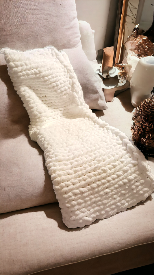 A white chunky knit blanket draped over the back of a beige sofa, with a textured pattern visible.