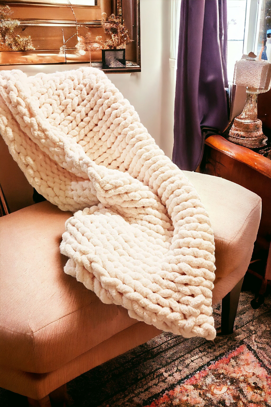 A chunky knit blanket in soft pink color is draped over the back of a beige armchair, with a lamp and wooden side table in the background.