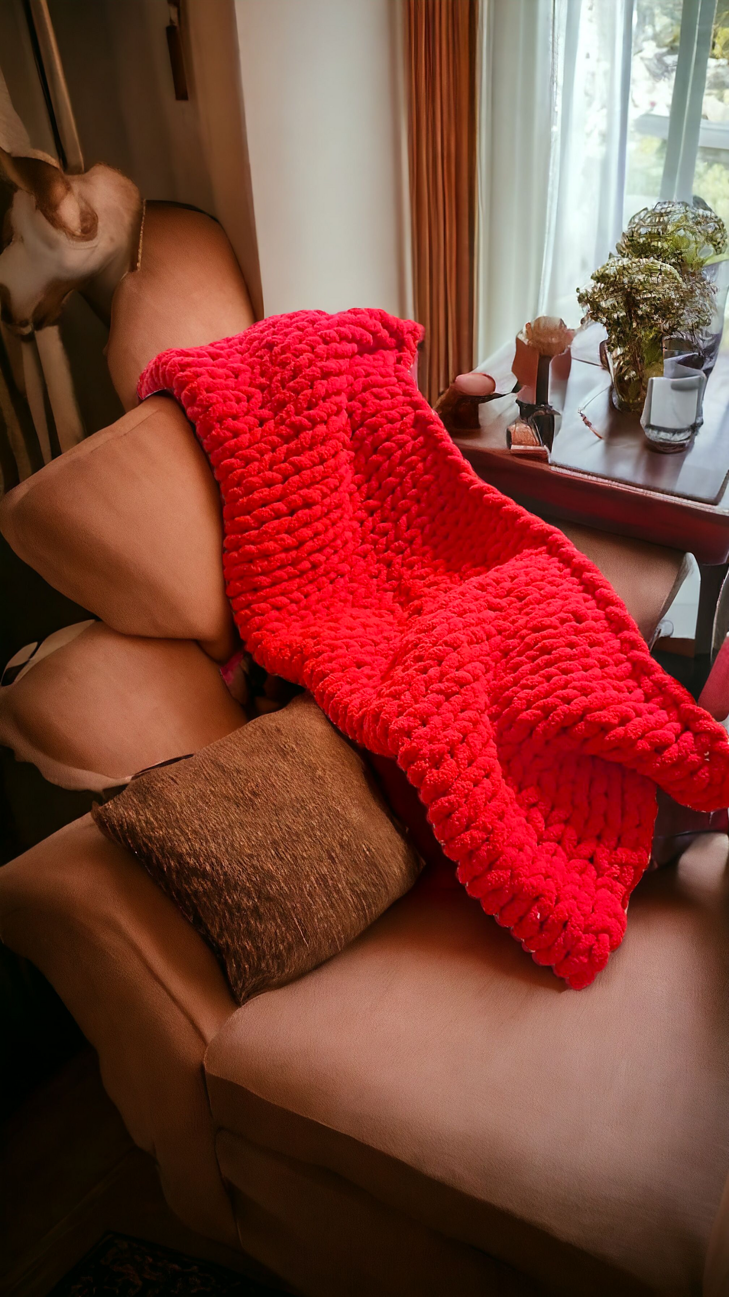 A red chunky knit blanket draped over a chair with pillows, set in a living room environment.