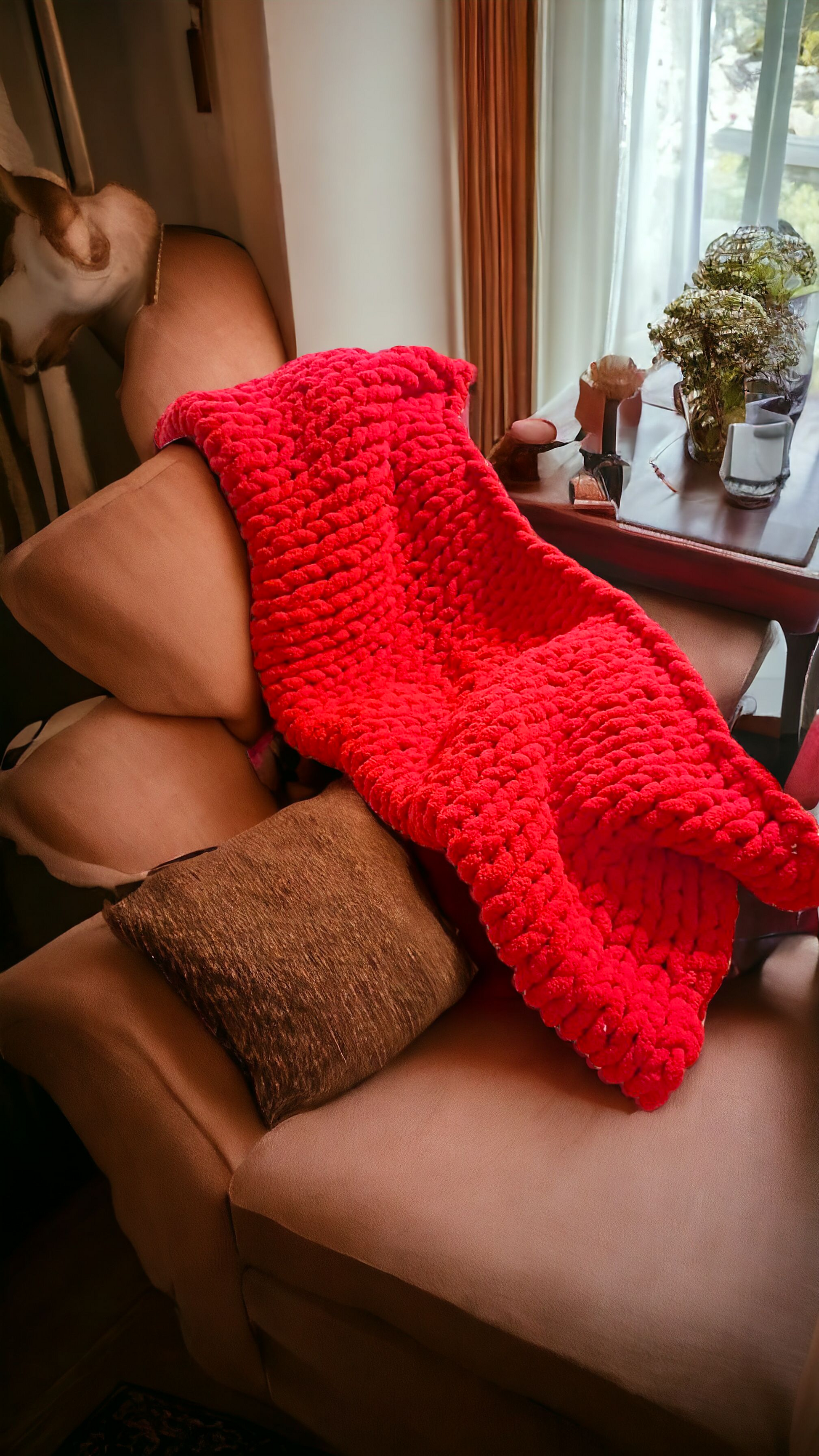 A red chunky knit blanket draped over a chair with pillows, set in a living room environment.