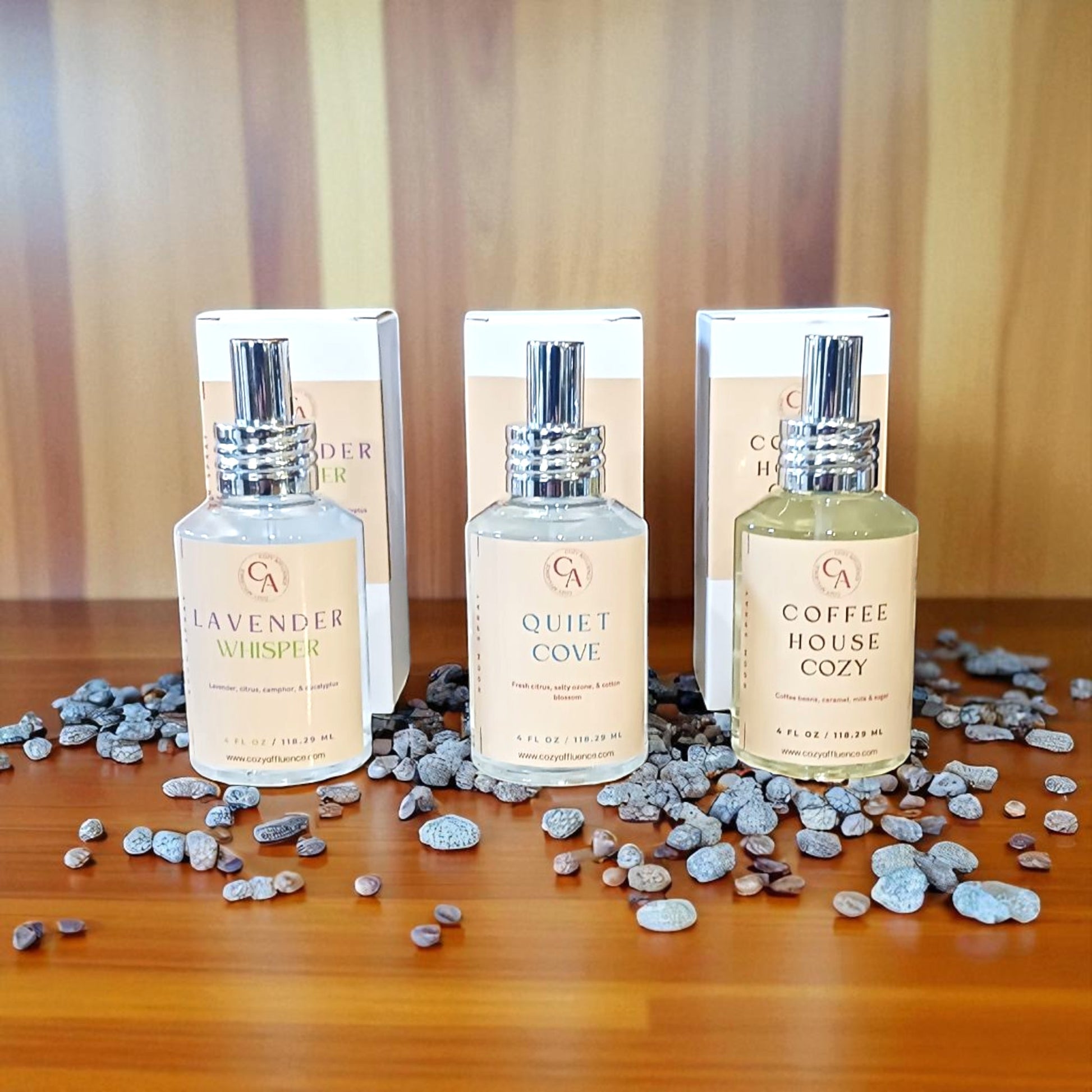 Three room spray bottles labeled 'Lavender Whisper', 'Quiet Cove', and 'Coffee House Cozy' on a wooden surface with decorative stones.