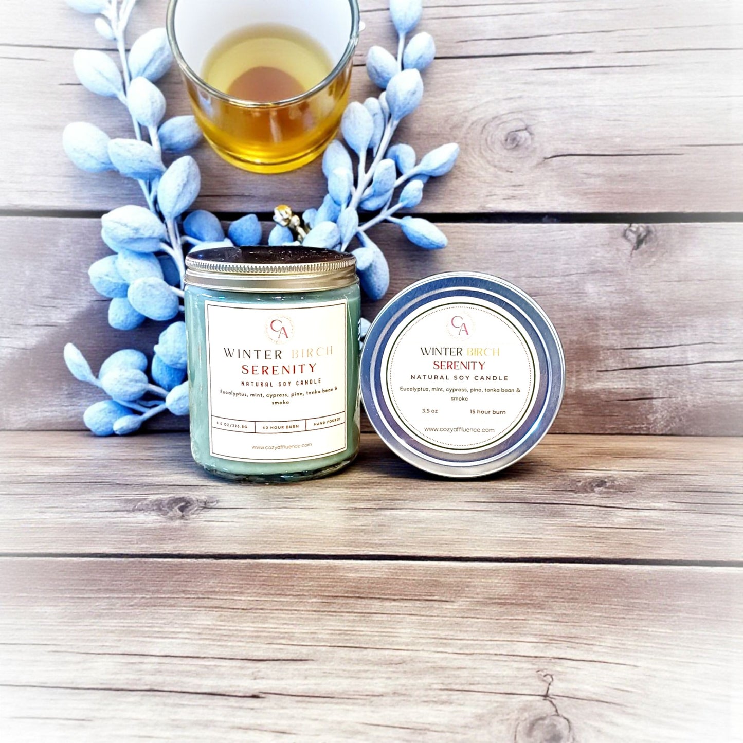 Two candles labeled 'Winter Serenity' on a wooden surface with a small glass of liquid and decorative branches.