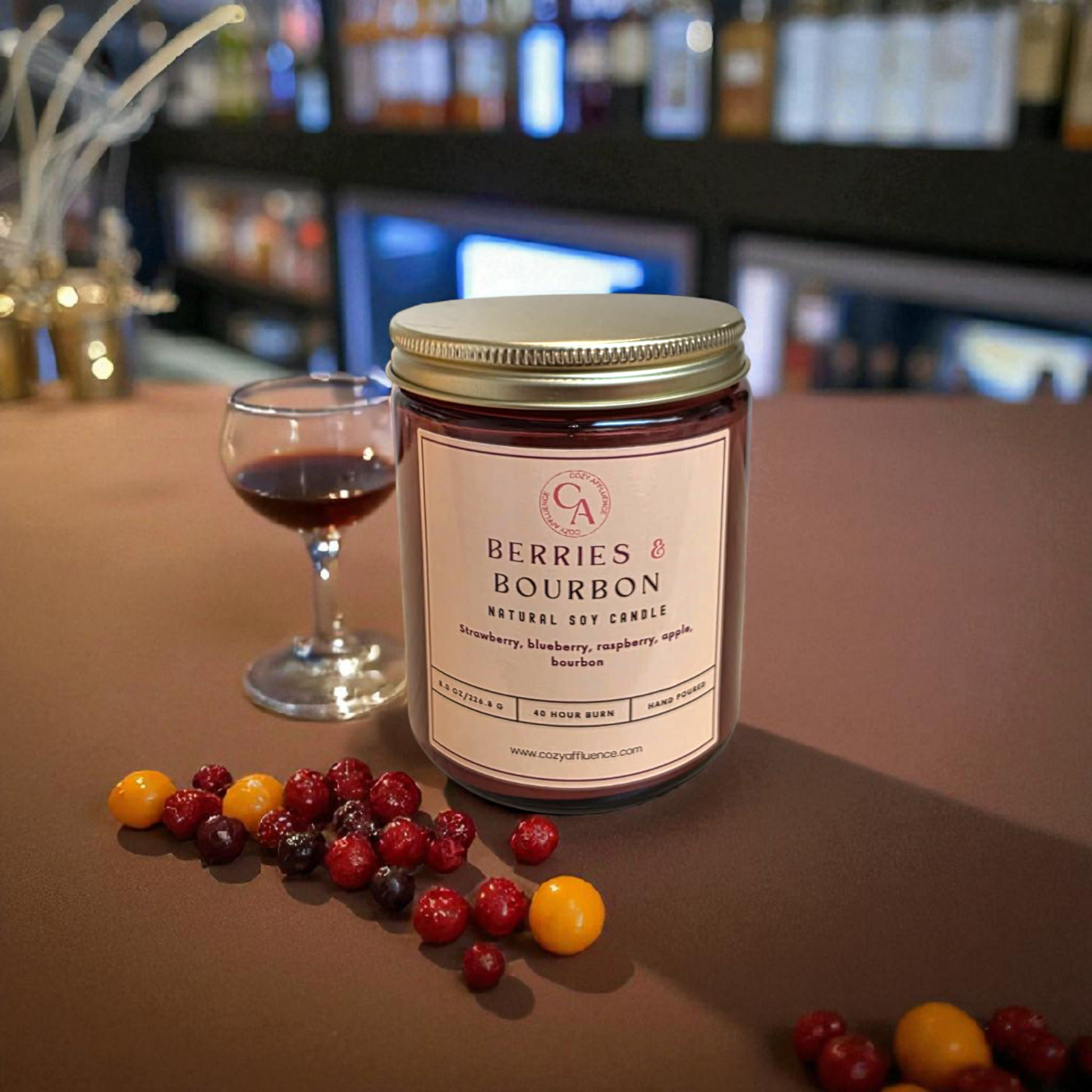 Berries and bourbon candle, sitting on top of a brown velvt table with fresh berries and bourbon in a glass
