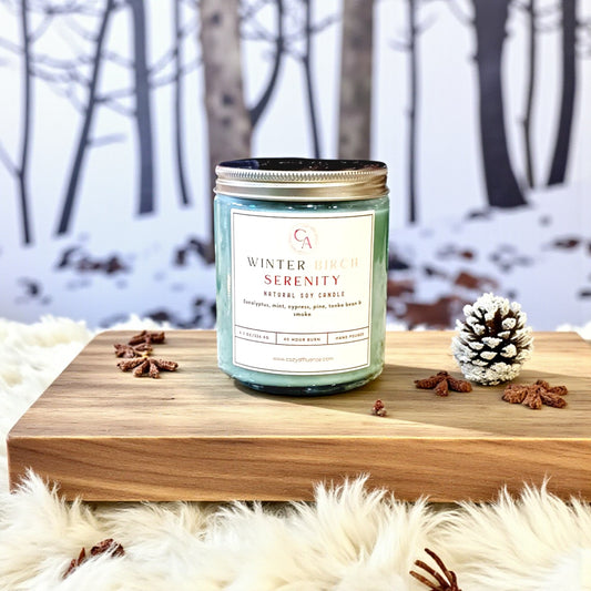Candle labeled 'Winter Serenity' on a wooden surface with a snowy forest background